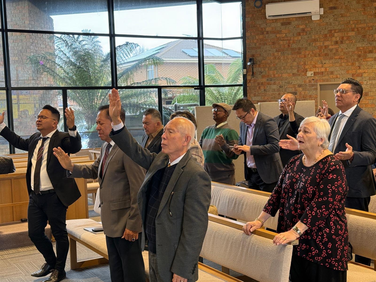 Gambar ini menampilkan momen penuh spiritual dalam ibadah pembukaan MUKERDA GPdI Australia 2025. Para jemaat, terdiri dari pria dan wanita berbagai usia dan latar belakang, berdiri dan mengangkat tangan dalam sikap penyembahan. Mereka mengenakan pakaian formal seperti jas dan kemeja; sebagian mengenakan dasi, menunjukkan kekhidmatan suasana. Di barisan depan, terlihat beberapa pemimpin gereja, termasuk pria lanjut usia dengan rambut putih dan jas abu gelap yang sebelumnya juga tampak dalam rapat. Seorang wanita dengan rambut putih dan baju hitam bercorak merah turut serta dalam penyembahan. Suasana ini berlangsung di dalam gereja berdinding bata ekspos berwarna coklat-oranye, dengan pencahayaan alami dari jendela kaca besar yang menampilkan pemandangan pepohonan di luar. Kehadiran para pemimpin dan jemaat yang antusias ini mencerminkan tema “Fervent Servants” dari MUKERDA tahun ini. Foto ini tidak hanya menangkap semangat pelayanan, tetapi juga memperlihatkan keragaman dan kekompakan dalam tubuh Kristus. Komposisi visual ini mendukung pengoptimalan SEO dengan kata kunci “pelayanan gereja di Australia” dan “ibadah pembukaan MUKERDA 2025”, menjadikan gambar ini kuat untuk pencarian visual yang relevan dengan aktivitas rohani dan organisasi GPdI.