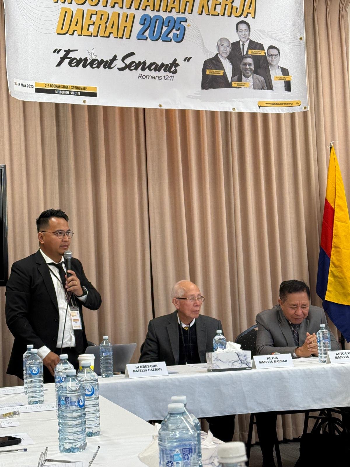 Gambar ini menampilkan momen penuh spiritual dalam ibadah pembukaan MUKERDA GPdI Australia 2025. Para jemaat, terdiri dari pria dan wanita berbagai usia dan latar belakang, berdiri dan mengangkat tangan dalam sikap penyembahan. Mereka mengenakan pakaian formal seperti jas dan kemeja; sebagian mengenakan dasi, menunjukkan kekhidmatan suasana. Di barisan depan, terlihat beberapa pemimpin gereja, termasuk pria lanjut usia dengan rambut putih dan jas abu gelap yang sebelumnya juga tampak dalam rapat. Seorang wanita dengan rambut putih dan baju hitam bercorak merah turut serta dalam penyembahan. Suasana ini berlangsung di dalam gereja berdinding bata ekspos berwarna coklat-oranye, dengan pencahayaan alami dari jendela kaca besar yang menampilkan pemandangan pepohonan di luar. Kehadiran para pemimpin dan jemaat yang antusias ini mencerminkan tema “Fervent Servants” dari MUKERDA tahun ini. Foto ini tidak hanya menangkap semangat pelayanan, tetapi juga memperlihatkan keragaman dan kekompakan dalam tubuh Kristus.