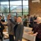 Pada gambar ini terlihat suasana formal Musyawarah Kerja Daerah (MUKERDA) GPdI Australia 2025 yang diselenggarakan di Melbourne. Seorang pria mengenakan jas hitam dan dasi sedang berdiri dengan memegang mikrofon, tampak sedang menyampaikan laporan atau arahan. Di meja depan, terdapat dua pria lanjut usia duduk berdampingan, masing-masing dengan label “Sekretaris Majelis Daerah” dan “Ketua Majelis Daerah”. Mereka mengenakan jas formal berwarna abu-abu dan hitam. Meja dipenuhi botol air mineral, tisu, dan dokumen. Latar belakang ruangan dihiasi tirai krem polos serta bendera merah-kuning-biru di sisi kanan gambar, yang menandakan suasana nasional dan spiritual. Di bagian atas foto, terdapat spanduk besar dengan latar putih bertuliskan "Musyawarah Kerja Daerah 2025" dengan tipografi biru-kuning dan tagline “Fervent Servants” mengacu pada Roma 12:11. Teks tambahan menyebutkan tanggal kegiatan, yakni 16–18 Mei 2025, berlokasi di 2–6 Boonah Street, Springvale, Melbourne, VIC. Terdapat pula ilustrasi lima tokoh GPdI berpakaian jas lengkap, memberikan kesan profesional dan rohani.