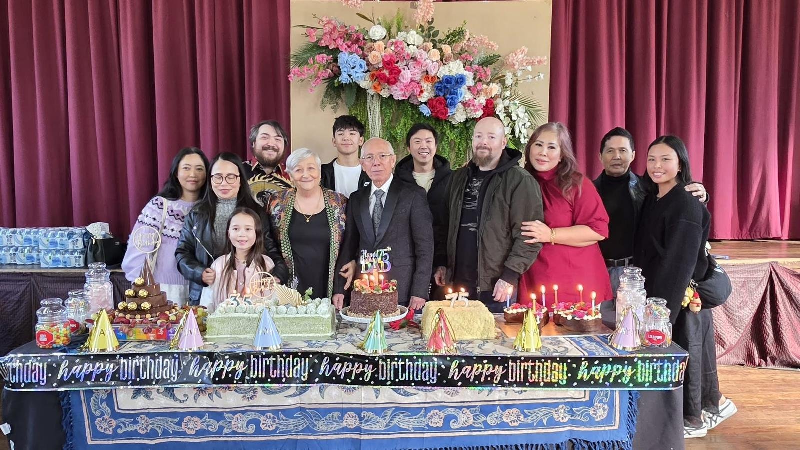 Foto perayaan ulang tahun ke-75 Ps Max Meiruntu bersama keluarga besar dan jemaat GPdI Australia yang berlangsung di Hall GPdI Sydney, Australia. Terlihat Ps Max Meiruntu berdiri di tengah mengenakan jas hitam dan dasi formal, didampingi istri dan anak-cucu serta para jemaat yang mengenakan pakaian kasual rapi dan berwarna cerah. Meja depan dihiasi dengan dekorasi kue ulang tahun, termasuk kue dengan topper bertuliskan "Happy 75 B’Day", serta dekorasi ulang tahun warna-warni seperti topi kerucut, lilin, dan tulisan "happy birthday" yang dicetak berulang kali pada banner hitam dengan font putih dan aksen warna neon. Background panggung menampilkan tirai merah marun dan dekorasi bunga segar bernuansa merah muda, biru, dan putih. Gambar ini mencerminkan suasana kekeluargaan dan sukacita dalam merayakan dedikasi panjang Ps Max Meiruntu sebagai pemimpin spiritual GPdI Australia, khususnya di GPdI Sydney selama lebih dari 35 tahun.