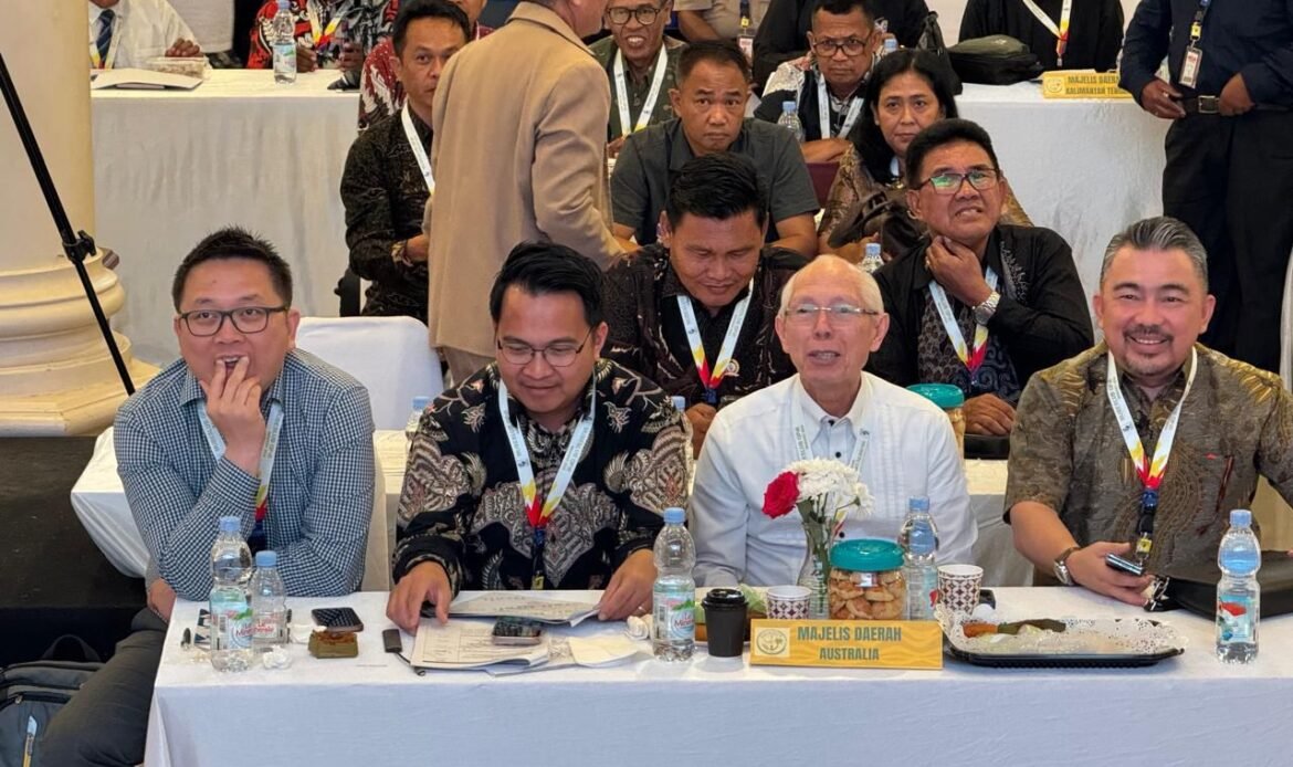 Foto delegasi Majelis Daerah GPdI Australia dalam acara Musyawarah Besar Luar Biasa (Mubeslub) GPdI 2025 di Kota Manado, Sulawesi Utara. Tampak deretan peserta duduk di meja panjang berlapis kain putih dengan botol air mineral, kopi, snack, dan vas bunga berisi hiasan mawar merah serta putih di tengah meja. Di bagian depan meja terdapat papan nama kuning bertuliskan “MAJELIS DAERAH AUSTRALIA” dengan huruf kapital berwarna hitam. Para peserta GPdI Australia yang duduk di barisan depan mengenakan kemeja formal, batik, serta jas, lengkap dengan tanda pengenal lanyard berwarna putih dengan corak pelangi. Suasana acara terlihat penuh khidmat, dengan latar belakang peserta lainnya yang mengenakan pakaian batik serta busana formal hitam, serta dekorasi ruangan rapih khas pertemuan gerejawi resmi. Foto ini mendokumentasikan keterlibatan GPdI Australia dalam sidang besar GPdI yang menjadi tonggak sejarah pelayanan di Manado tahun 2025.