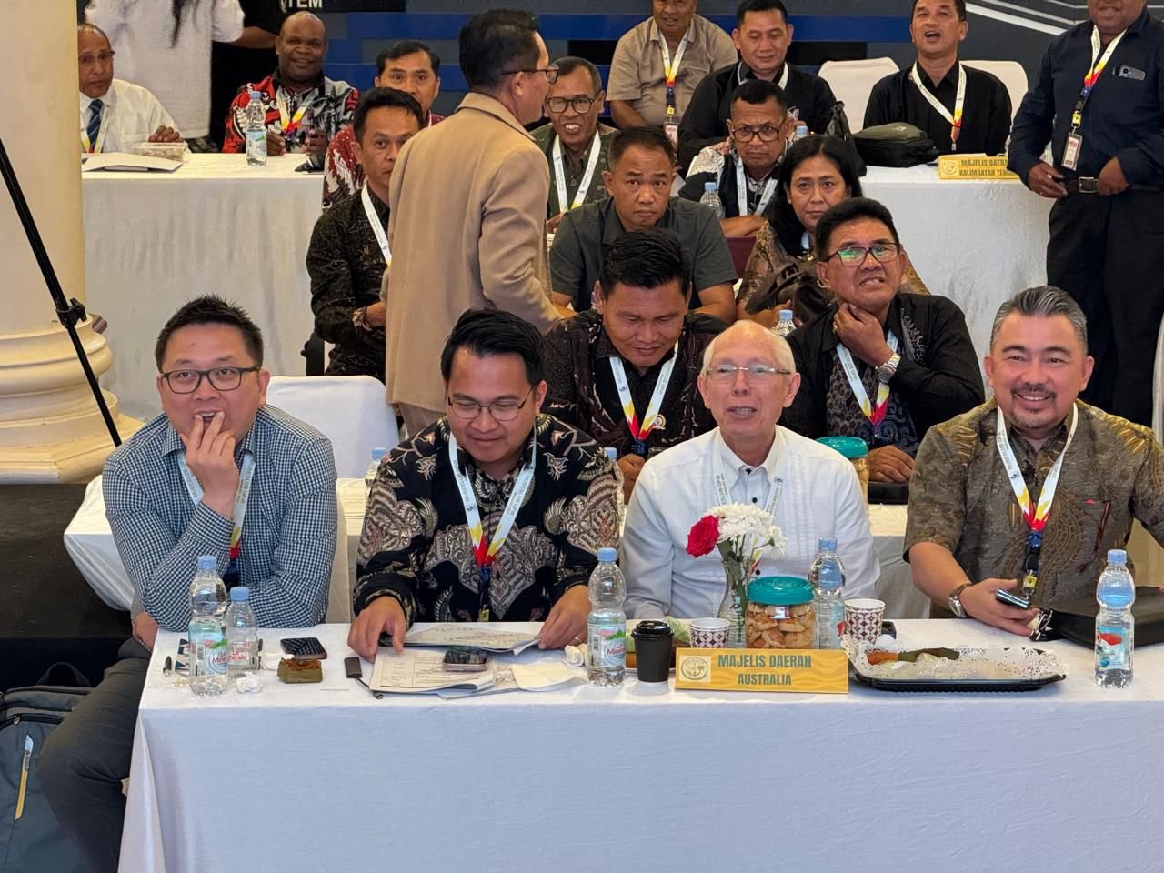 Foto delegasi Majelis Daerah GPdI Australia dalam acara Musyawarah Besar Luar Biasa (Mubeslub) GPdI 2025 di Kota Manado, Sulawesi Utara. Tampak deretan peserta duduk di meja panjang berlapis kain putih dengan botol air mineral, kopi, snack, dan vas bunga berisi hiasan mawar merah serta putih di tengah meja. Di bagian depan meja terdapat papan nama kuning bertuliskan “MAJELIS DAERAH AUSTRALIA” dengan huruf kapital berwarna hitam. Para peserta GPdI Australia yang duduk di barisan depan mengenakan kemeja formal, batik, serta jas, lengkap dengan tanda pengenal lanyard berwarna putih dengan corak pelangi. Suasana acara terlihat penuh khidmat, dengan latar belakang peserta lainnya yang mengenakan pakaian batik serta busana formal hitam, serta dekorasi ruangan rapih khas pertemuan gerejawi resmi. Foto ini mendokumentasikan keterlibatan GPdI Australia dalam sidang besar GPdI yang menjadi tonggak sejarah pelayanan di Manado tahun 2025.