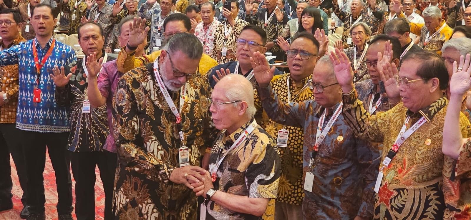 Foto suasana penuh khidmat dalam acara Rapat Kerja Nasional (Rakernas) PGPI 2025 di Hotel Novotel Mangga Dua Square, Jakarta. Tampak para pemimpin gereja dan gembala sidang mengenakan batik bernuansa cokelat, biru, dan emas, dengan lanyard resmi Rakernas tergantung di leher. Di tengah gambar, tampak momen pelantikan dan doa peneguhan bagi Pdt. Max Meiruntu sebagai Ketua PGPI Australia, di mana para hamba Tuhan menumpangkan tangan dan berdoa bersama. Gambar ini merefleksikan semangat kebersamaan dan tema “Berbeda-beda Tetapi Satu” yang diusung oleh Persekutuan Gereja-gereja Pentakosta Indonesia (PGPI) dalam Rakernas PGPI 2025.